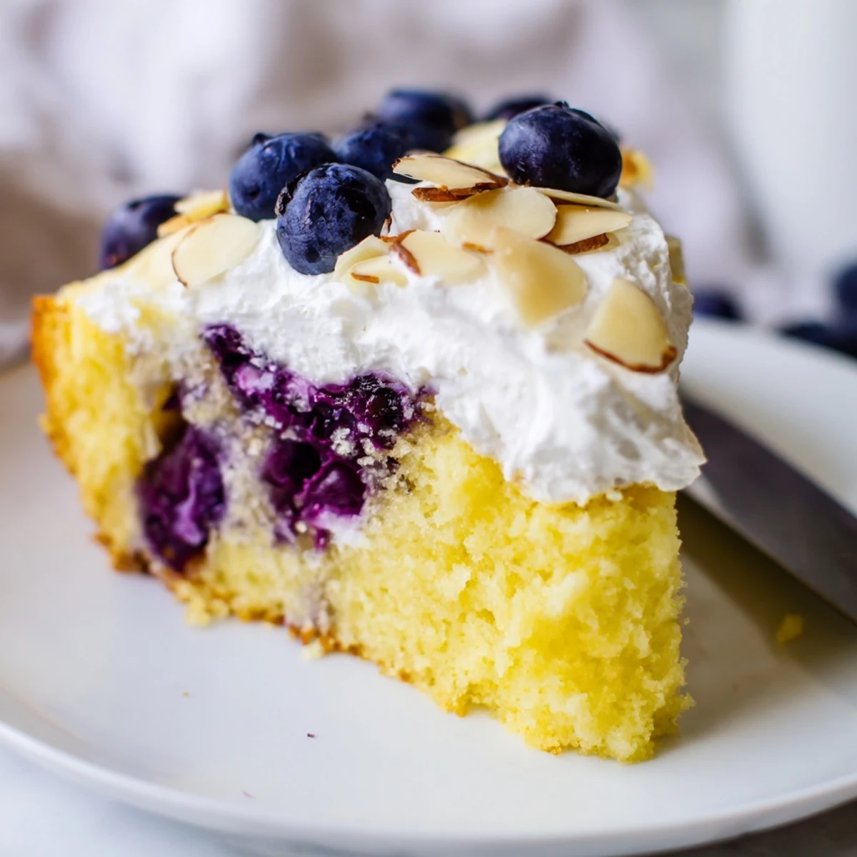 Generous square of almond-topped poke cake featuring bright lemon flavor and juicy blueberry pockets throughout